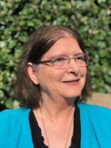Headshot photo of Mona Kaufmann in a blue top smiling with lush greenery behind her.